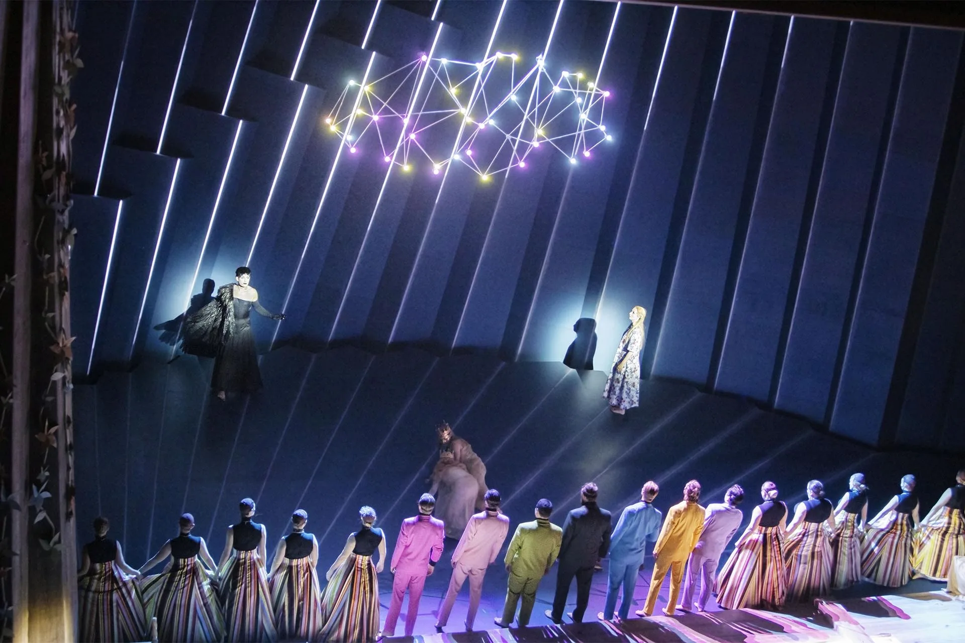 The chorus lined up across the front of the stage in striped coats, blue pixel lines and a warm constellation overhead