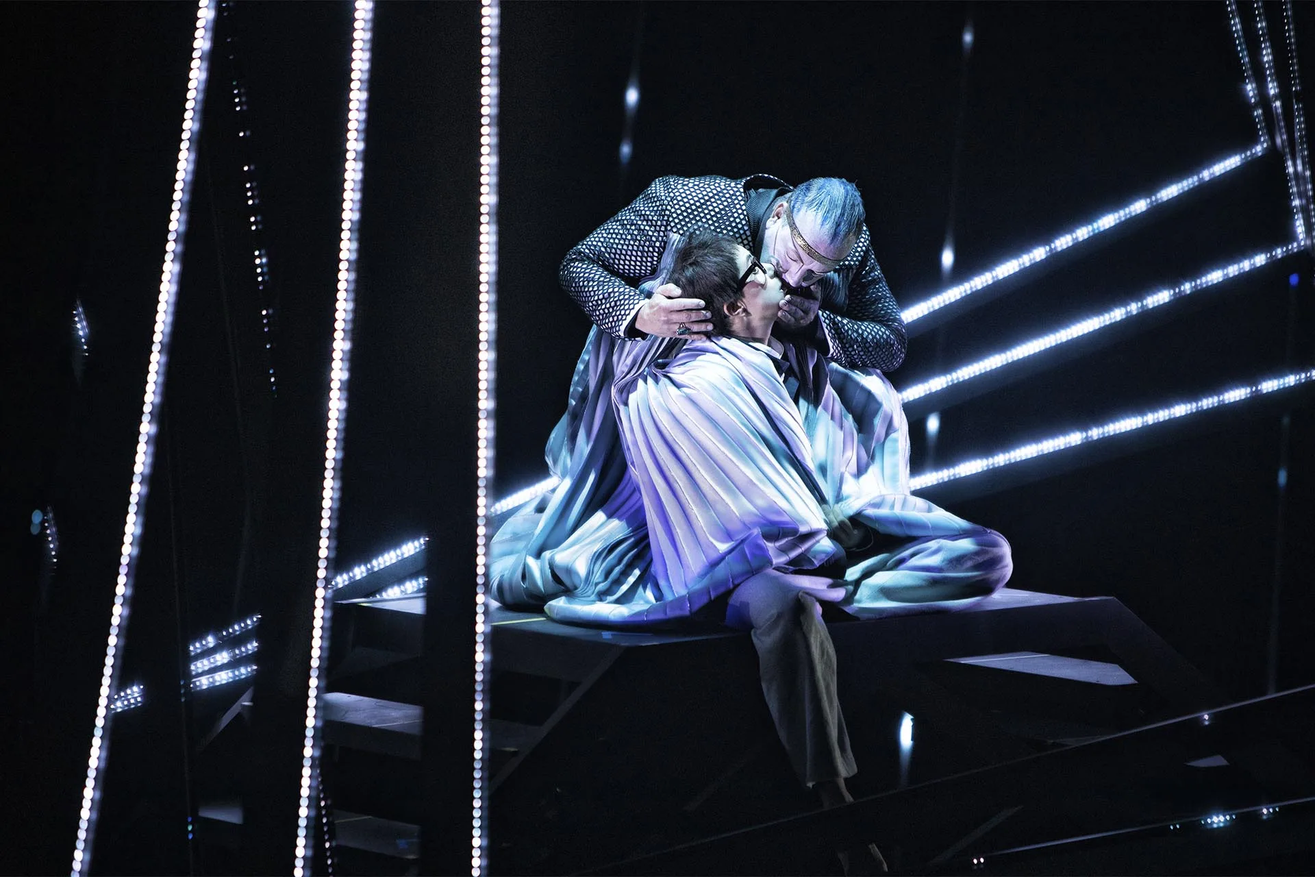 Two singers holding each other centre stage, tall vertical pixel strips running white on either side