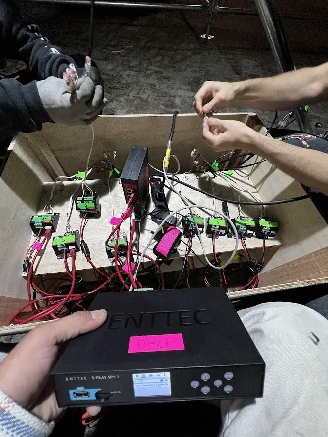 Hands wiring a cardboard box of green PLink injectors on site, with an ENTTEC S-PLAY SP-1 controller held up in front