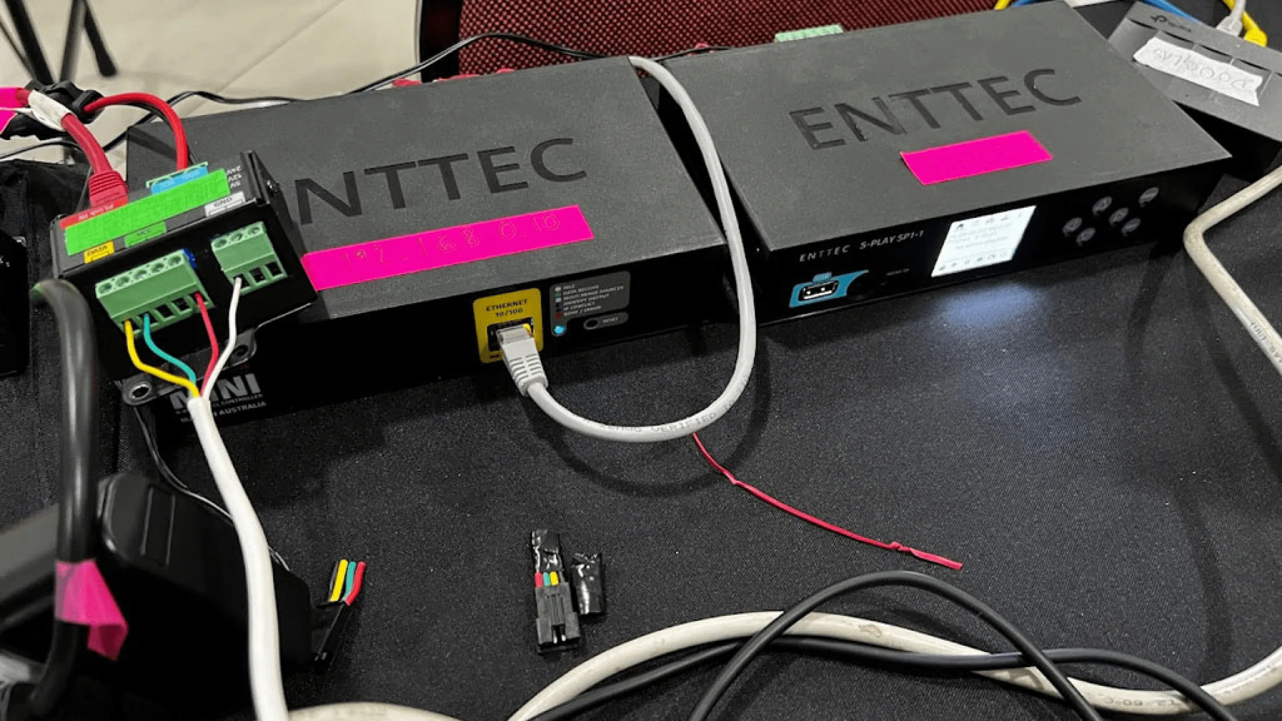 Two black ENTTEC controllers taped with pink labels on a desk, ethernet leads and red and black power wires running off them