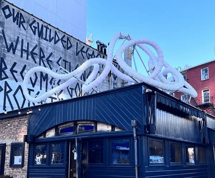 The unlit white sculpture mounted above a blue-painted corner diner in daylight, hand-painted mural text on the wall behind