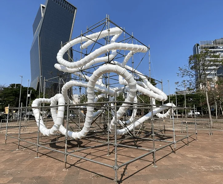 White unlit tubing in a scaffold cube on a paved plaza, glass office towers and trees behind in daylight