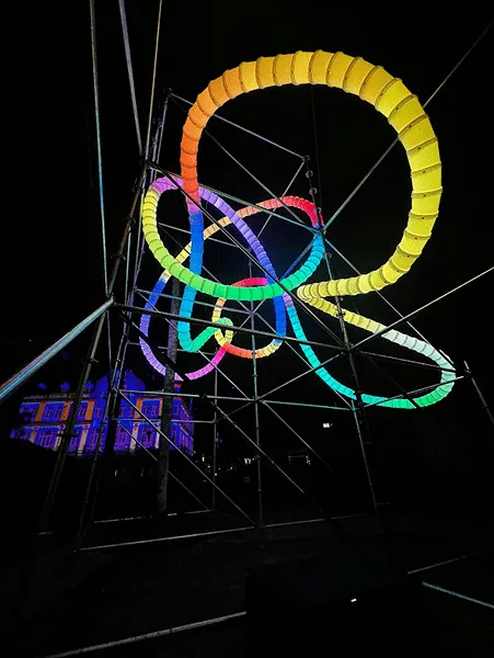 Loops lit yellow, green, blue and pink inside a black scaffold frame at night, guy wires crossing the dark background