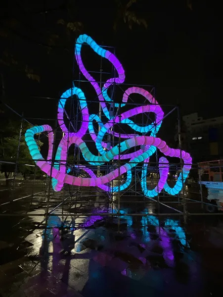 Blue, purple and cyan loops on a scaffold at night, the colour reflected in the wet paving below