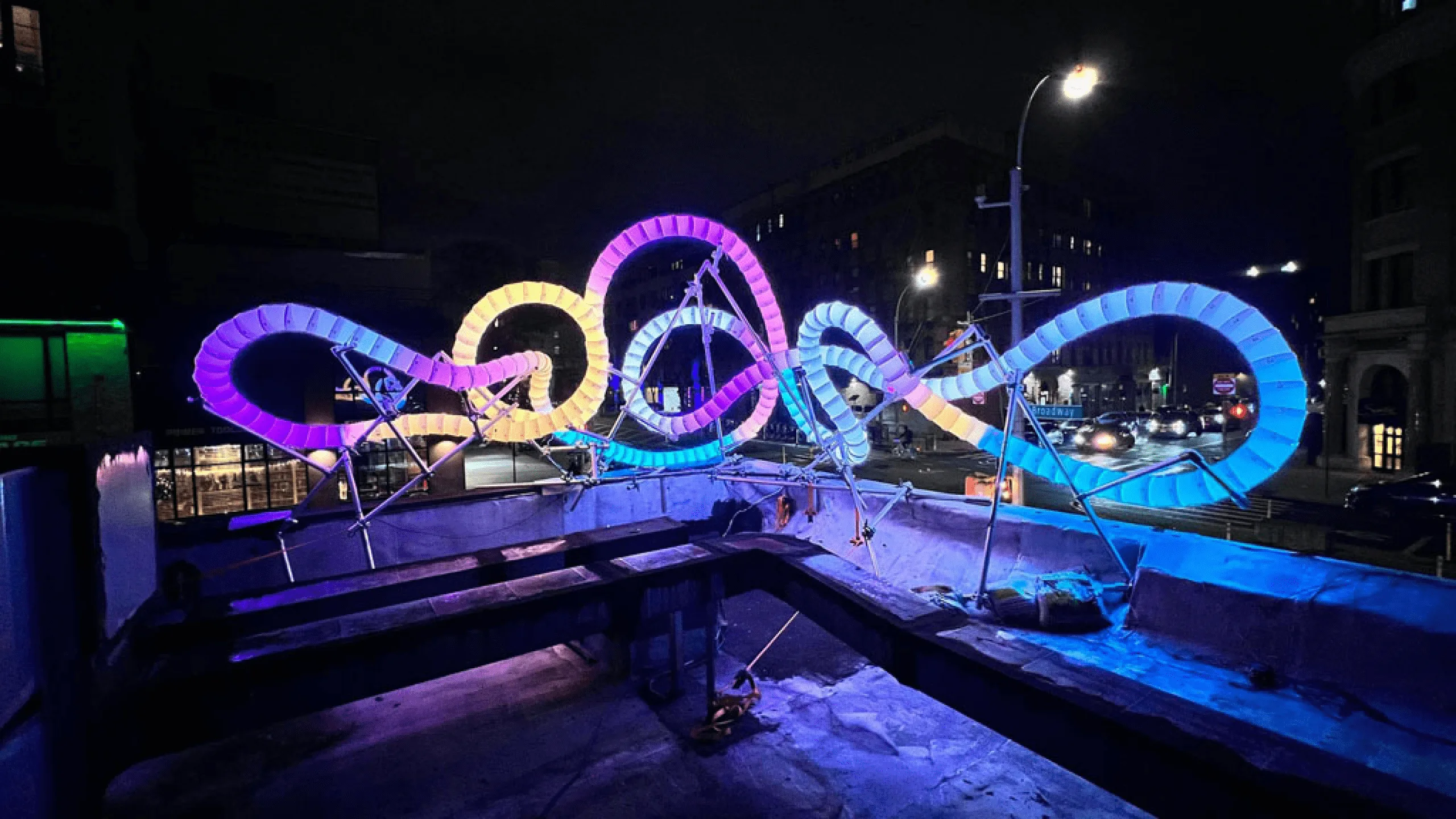 The sculpture glowing pink, cyan and blue on a raised deck beside a road at night, a streetlight flaring overhead