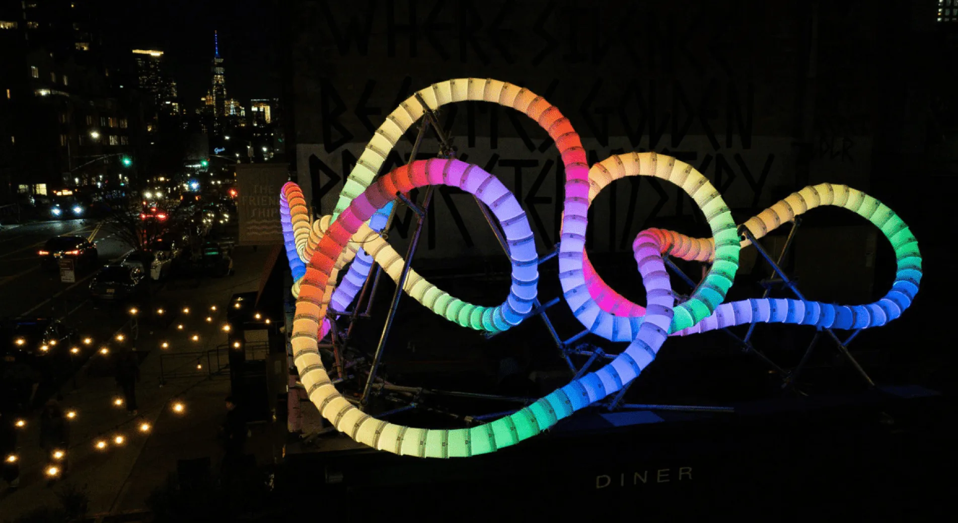 Rainbow-lit loops of the bucket sculpture arcing above a diner awning at night, city skyline and street traffic behind