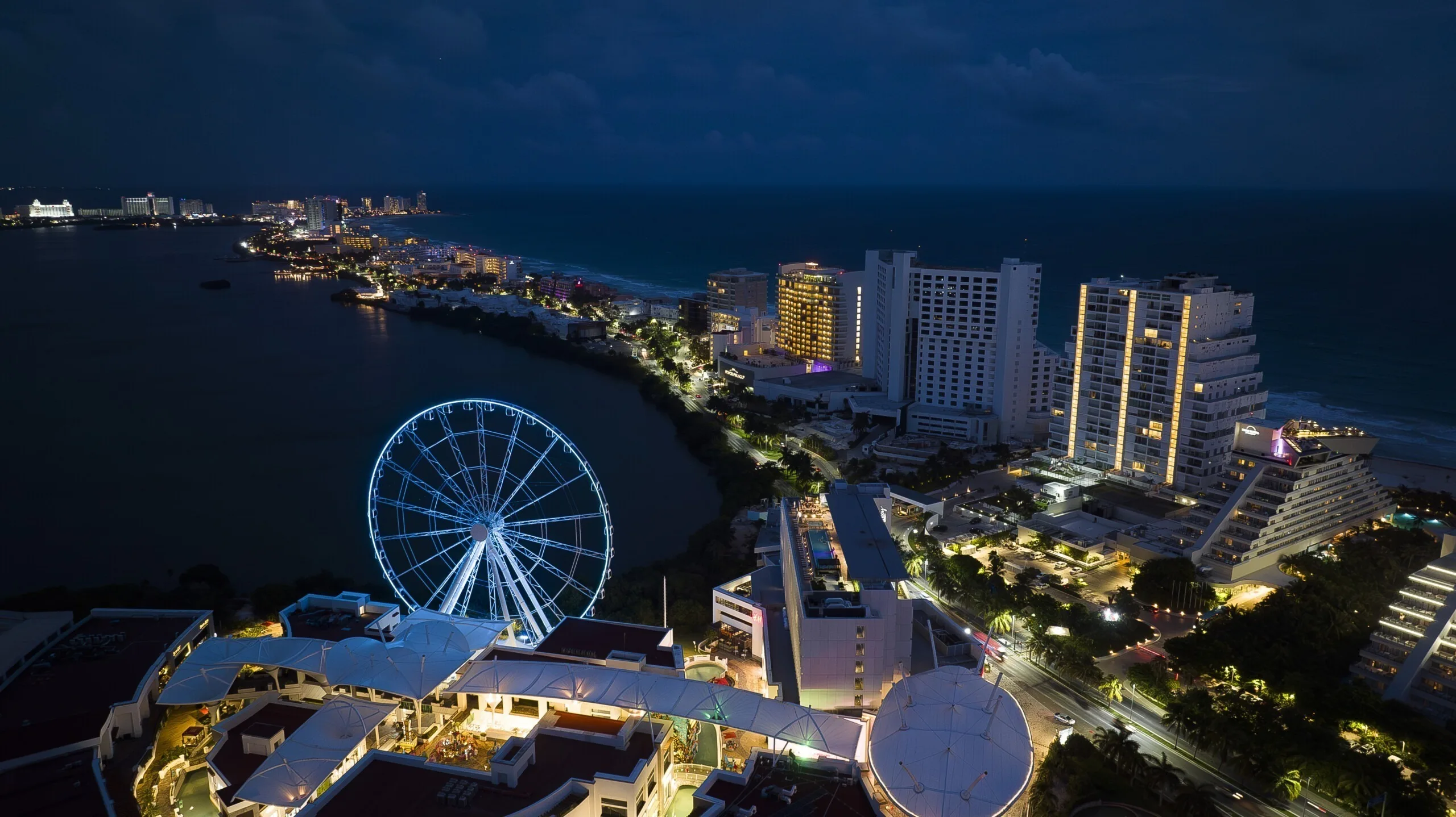 The wheel in cyan and white beside the lagoon, hotel towers lit along the shore to the right