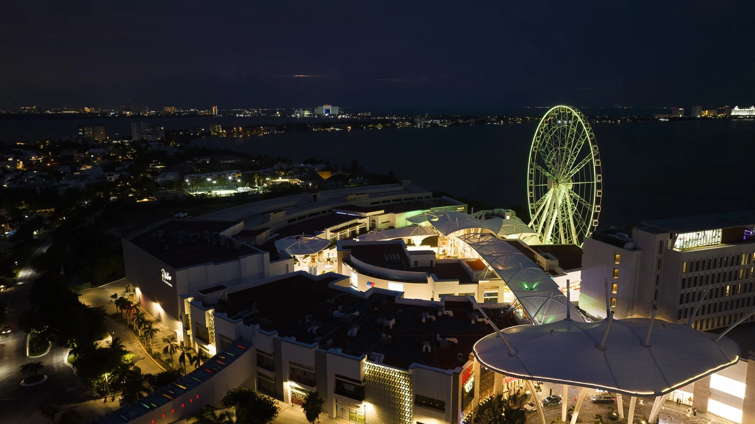 The wheel in white seen from the other side, the lit mall below it and city lights along the far shore