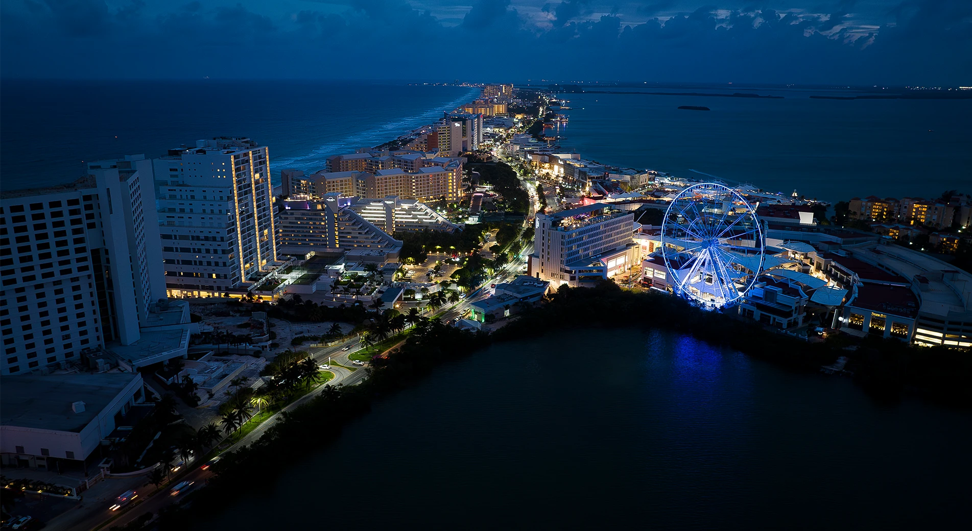 High aerial of the Cancun hotel strip after dark, the Sky Wheel lit blue above the mall on the right, the black lagoon in the foreground and lit hotels running away to the sea