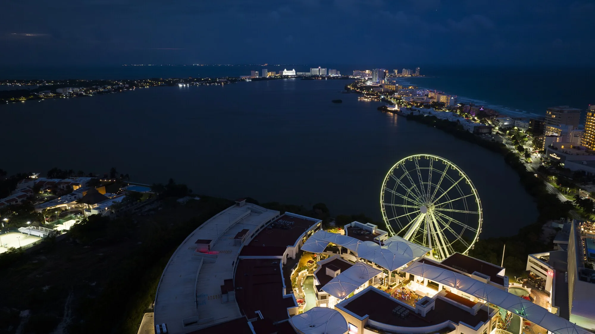 Dusk aerial of the wheel picked out in yellow and green light, the lagoon and the lit hotel strip stretching away behind it
