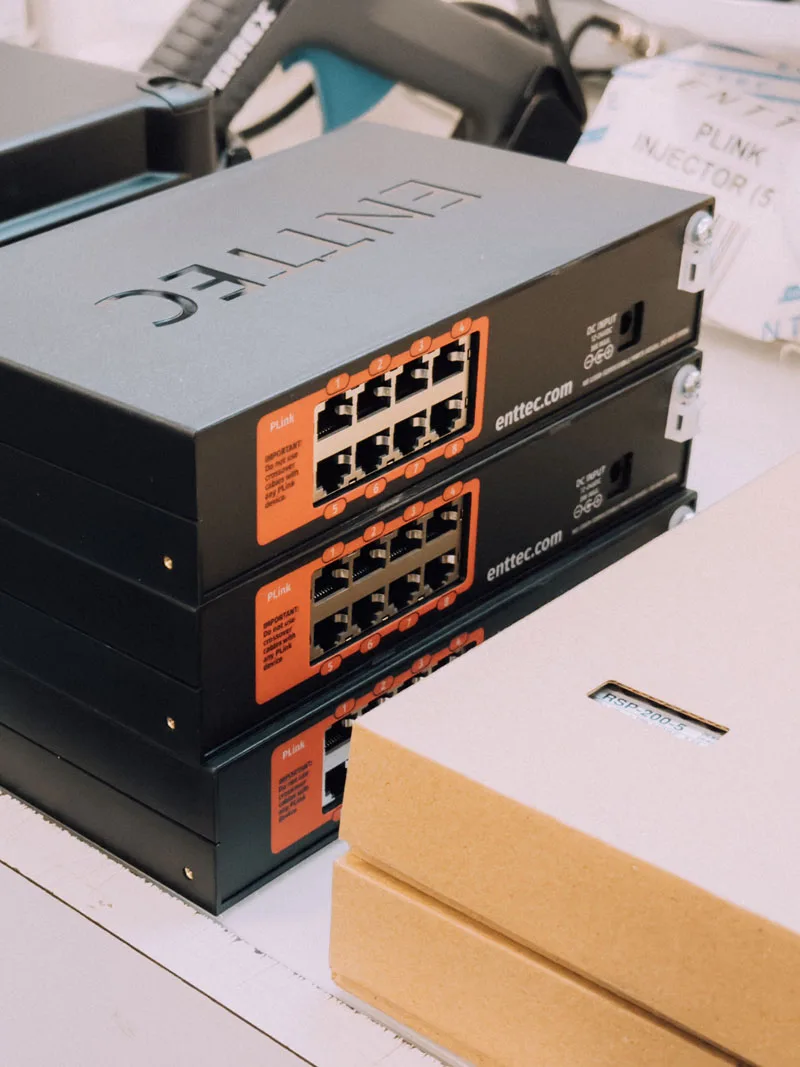 Two black ENTTEC controllers on a bench with their orange labelled output ports facing out, PLink injector cartons stacked behind