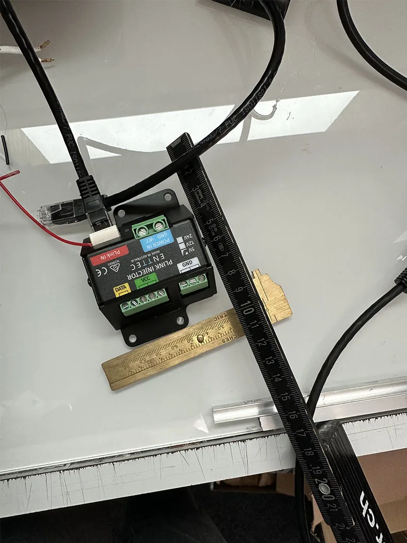A PLink injector lying on a white worktop with its cables run out, a brass ruler and a length of black conduit alongside