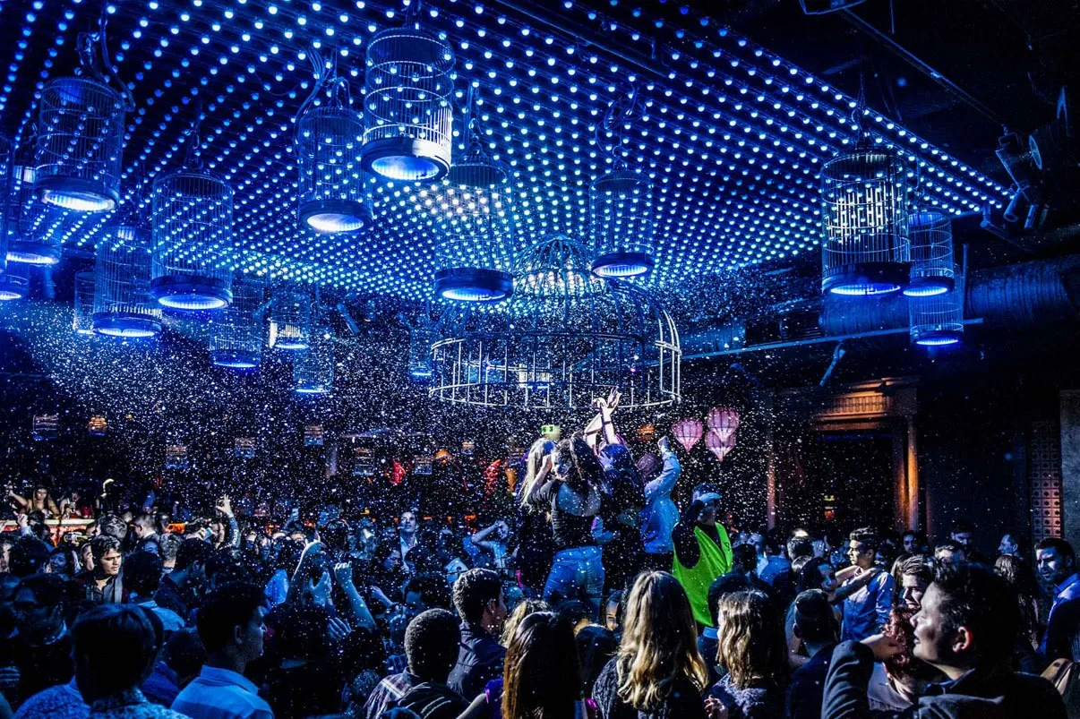 Wide view of the packed room, the whole ceiling of white dots glowing deep blue with confetti drifting down over the crowd
