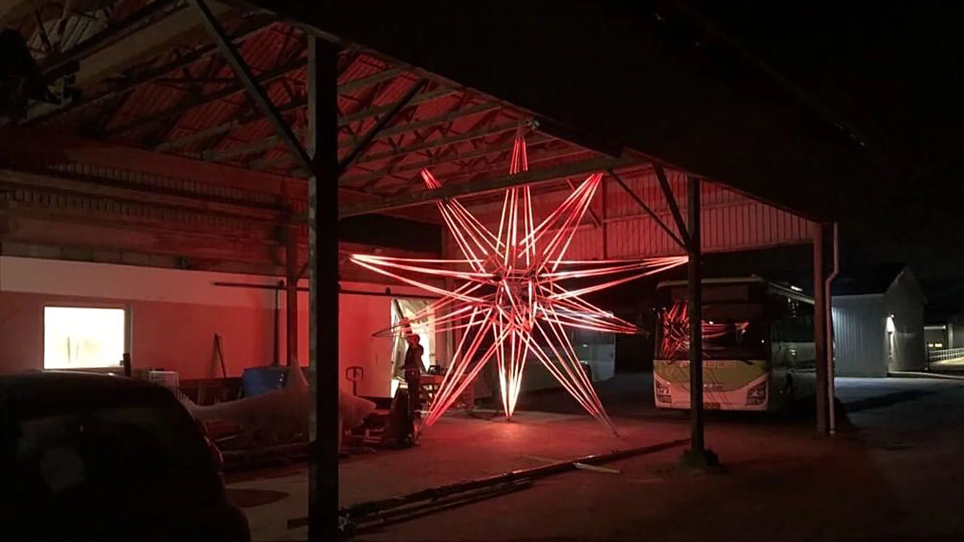 The star during the build inside a red lit depot, spokes glowing between the roof columns with a car and a coach parked either side