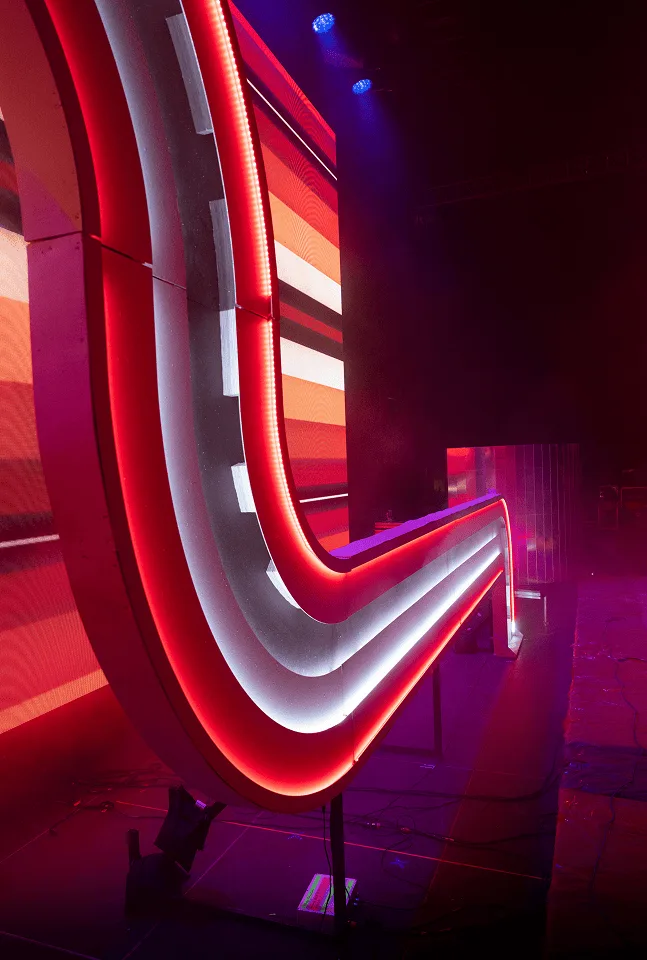 The arch from the side in red and white, its stacked strips doubling back on themselves against the video wall