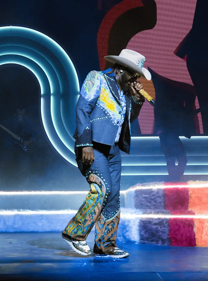 T-Pain in a white cowboy hat and studded blue jacket singing into the mic, the teal arch curving behind him
