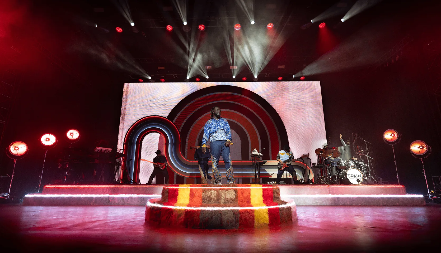 The stage from the crowd through heavy haze, red wash over the band and drum riser with the white screen behind