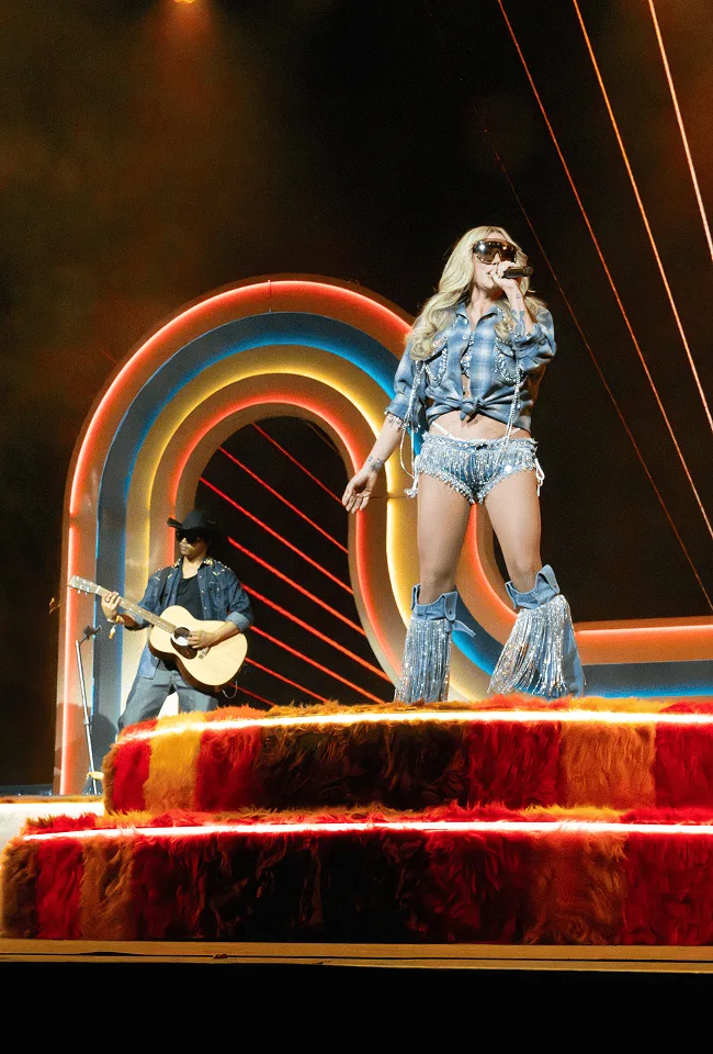 A guest singer in denim standing on the fur riser, a guitarist below her and the arch glowing warm behind