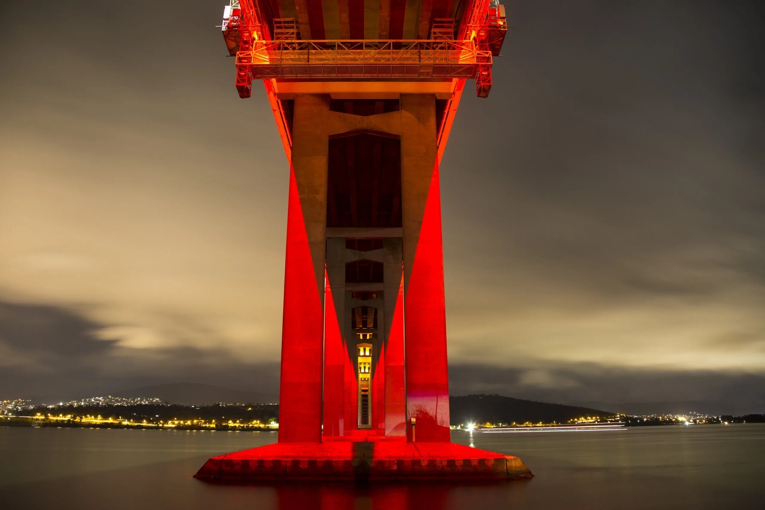 Close view up at the underside of the deck, red pylon against an olive gold sky and distant harbour lights
