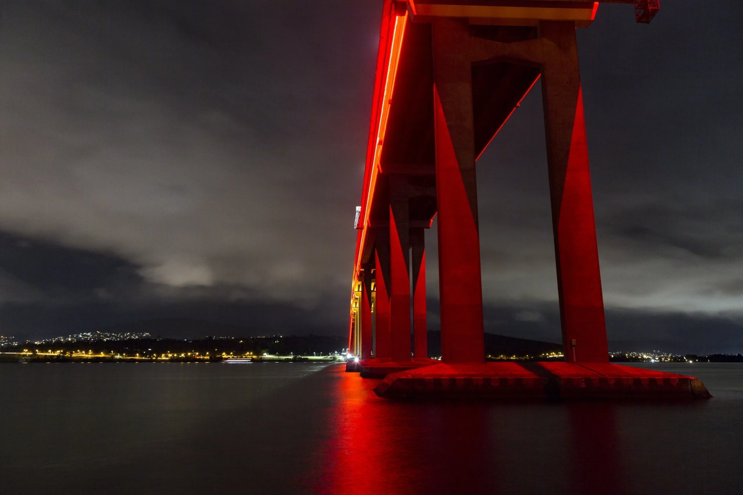 The same pylon from further back under dark cloud, red light spilling onto the water around its base