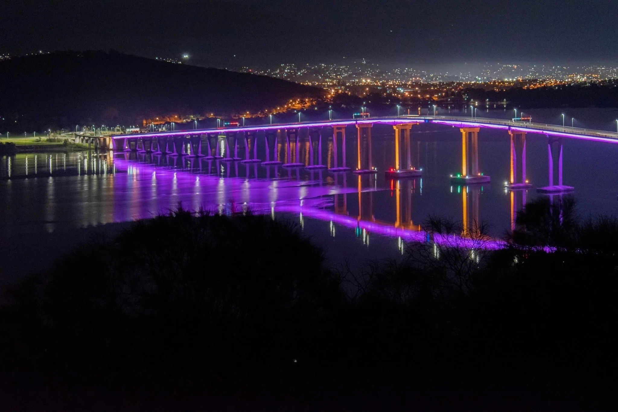 The whole span lit magenta and purple at night, hillside suburbs and city lights scattered behind it