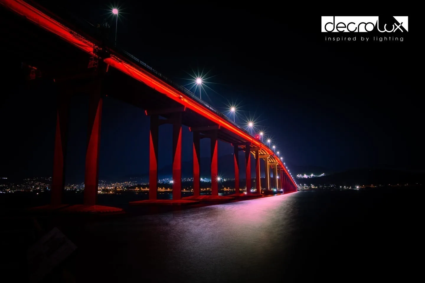 The same run of red piers with a pale pink pool of light on the water and streetlights flaring above the deck