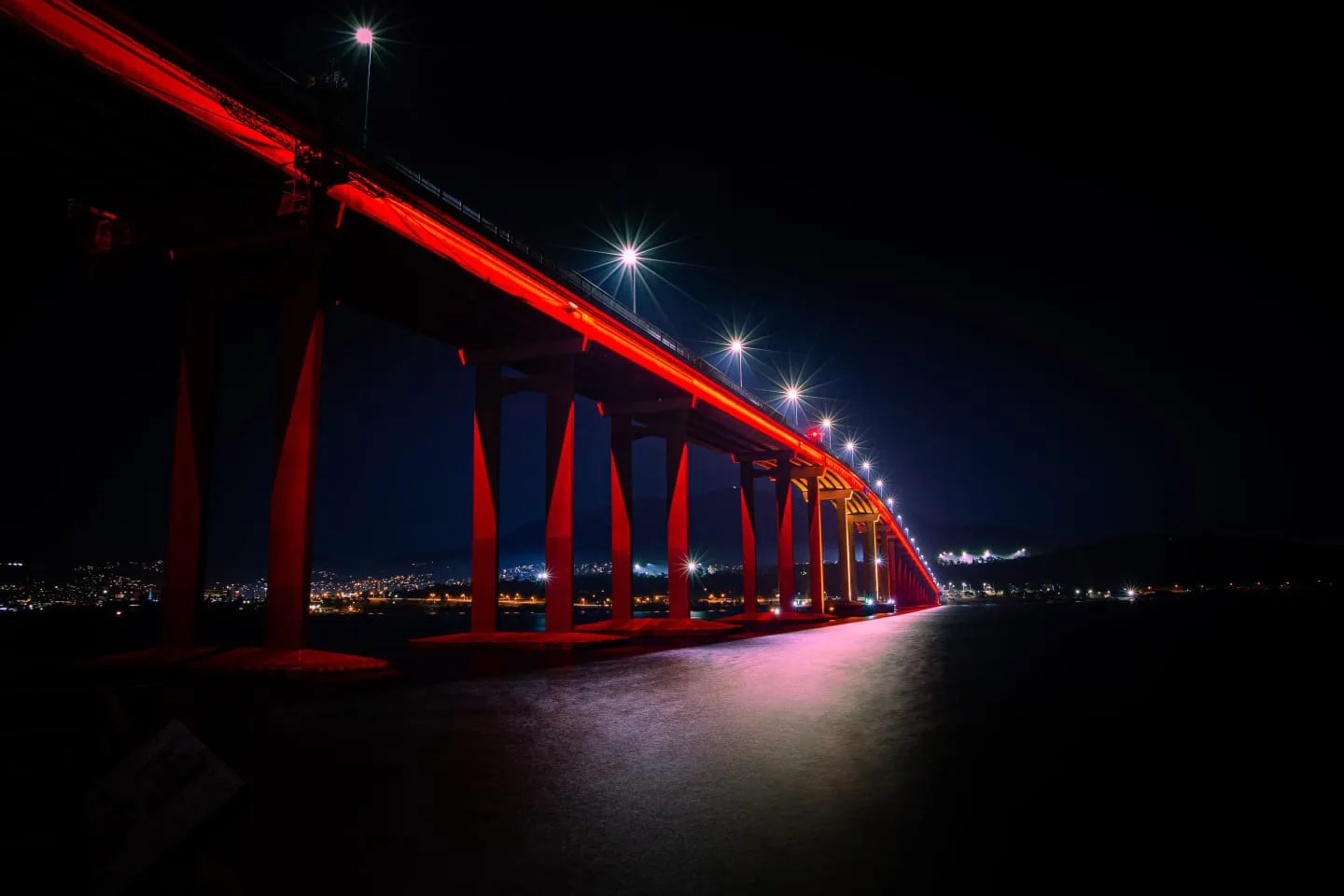Tighter, darker view along the red piers, the near columns in deep shadow and the city strung out on the far bank