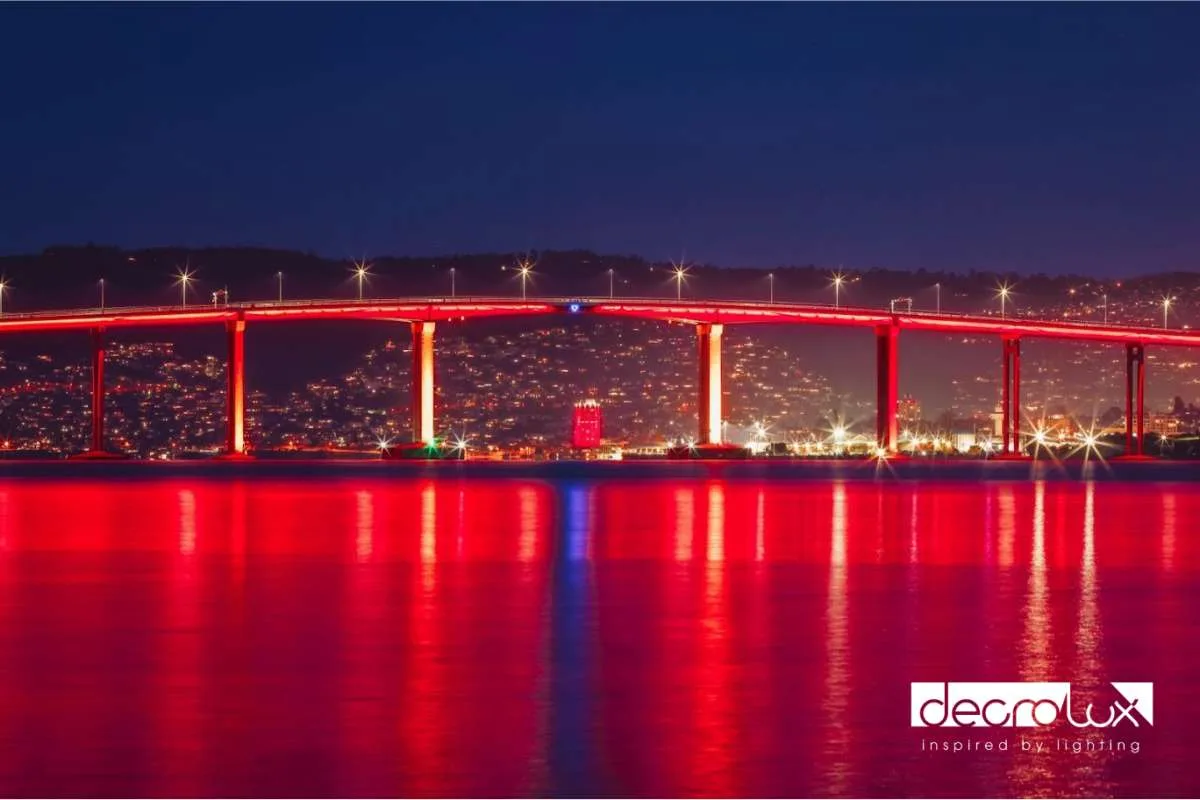 The span in red across the water at dusk, tall red reflections streaking toward the camera past the lit waterfront