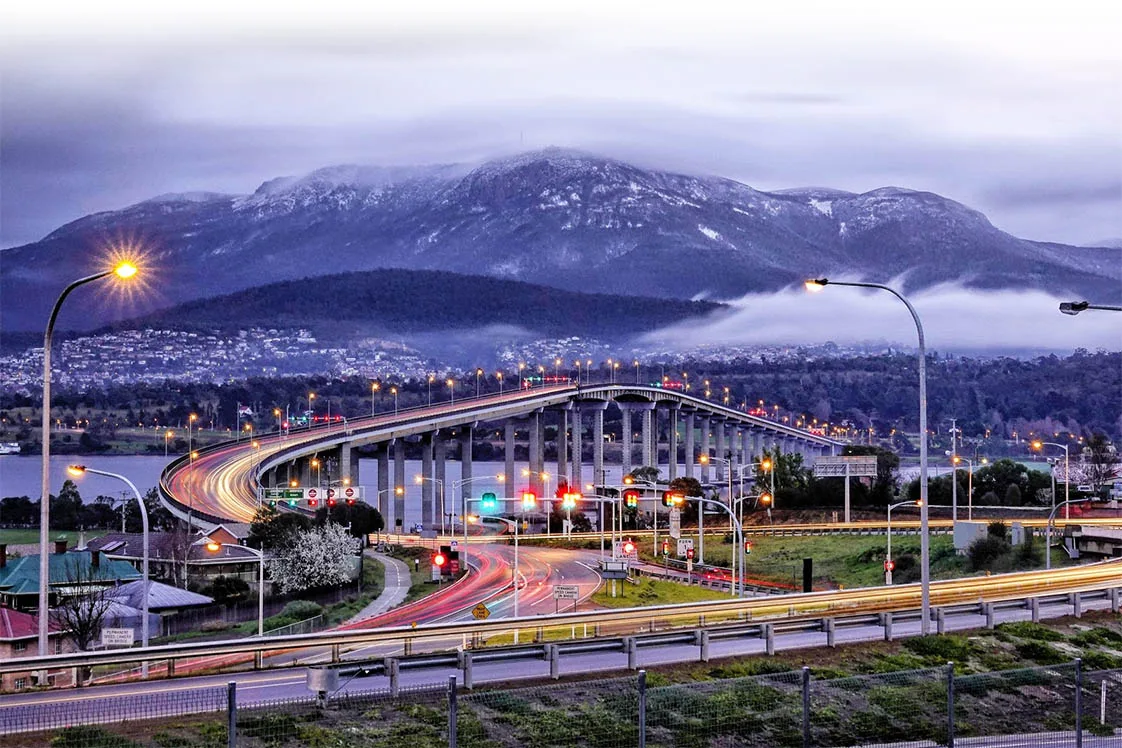 The bridge and its highway approach at dusk, car light trails curving beneath a snow-dusted Mount Wellington behind Hobart