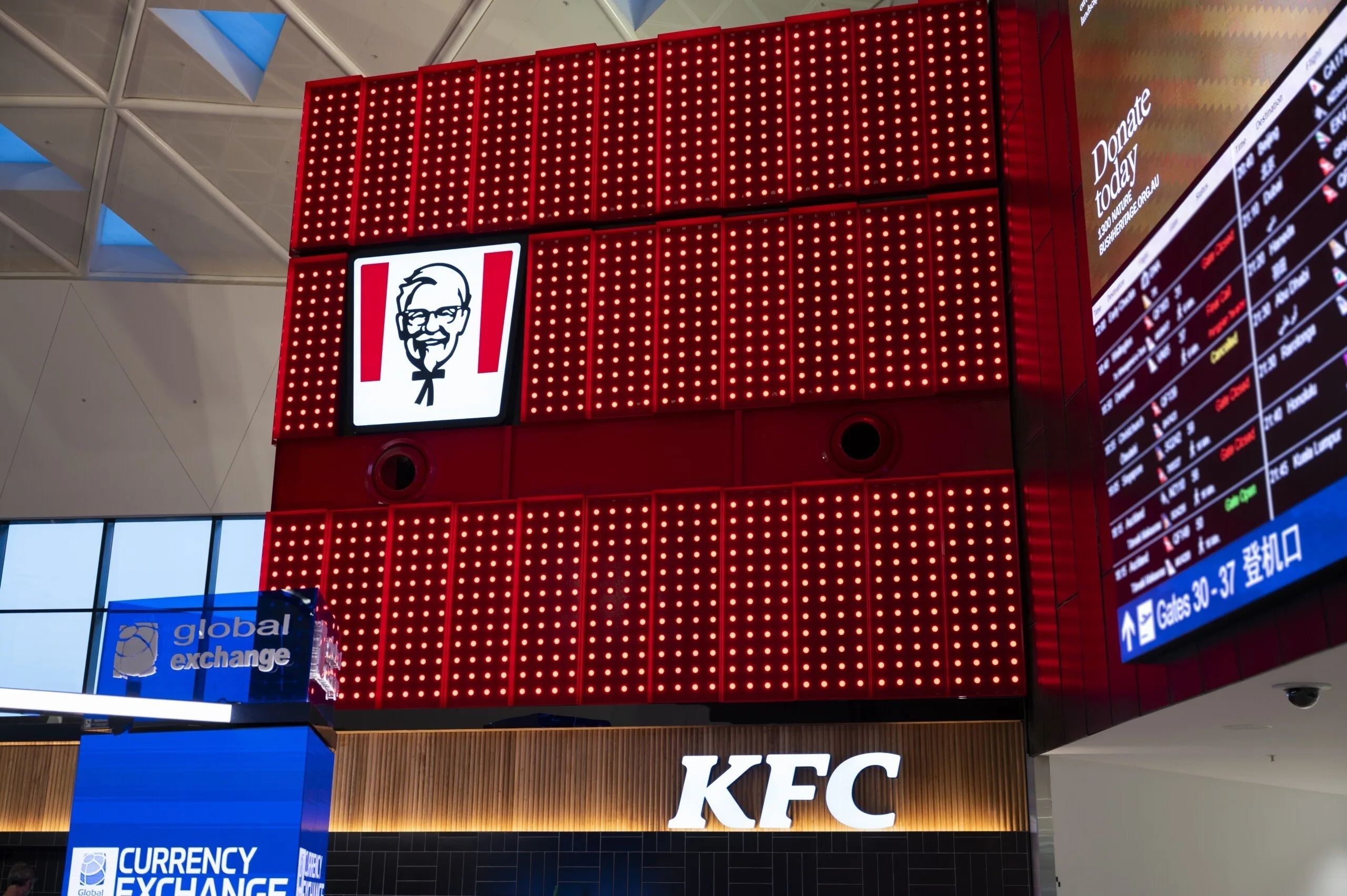 The box corner carrying the white KFC wordmark, flight information screens and a currency exchange booth below
