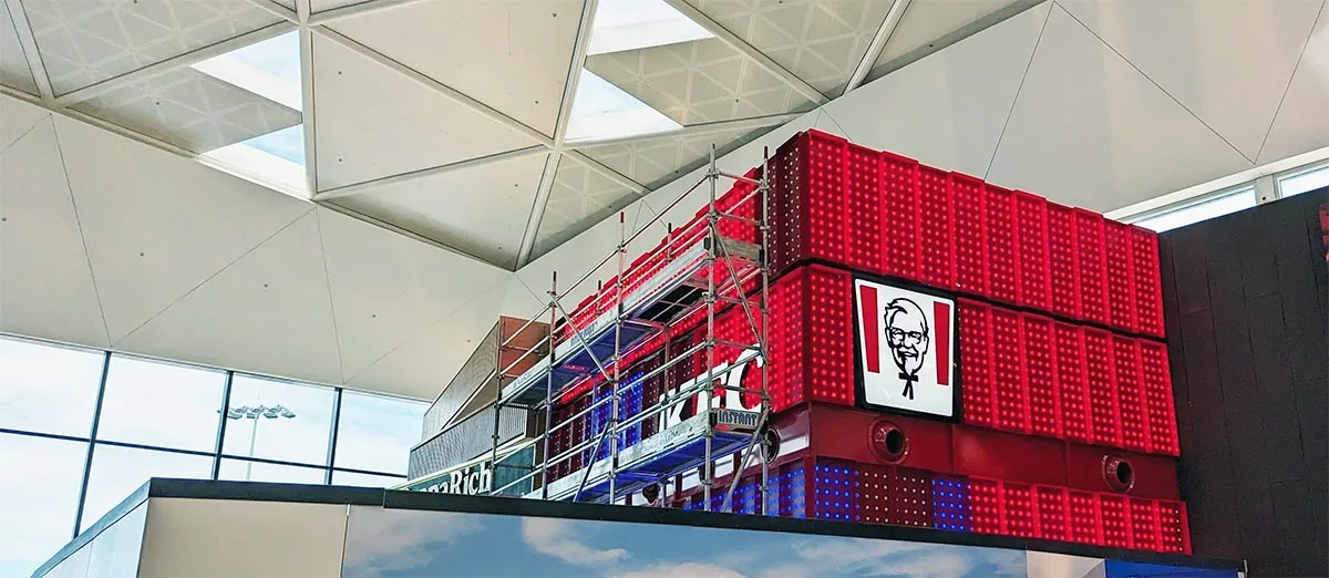 Scaffolding still standing against the unfinished side of the box while the completed red face and Colonel Sanders panel glow above the balcony
