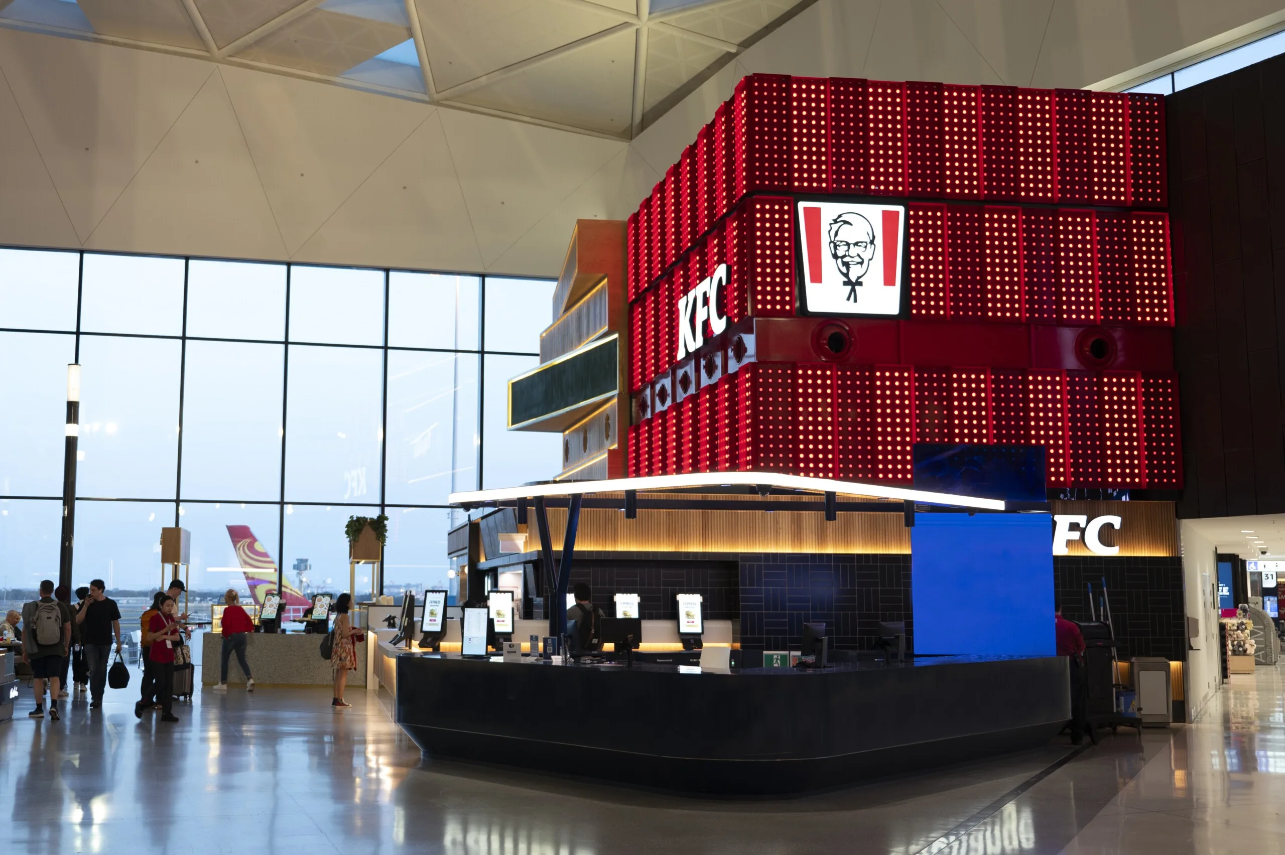 The KFC counter at Sydney Airport beneath its giant red pixel-dot box, travellers passing the glazed wall with the tarmac beyond