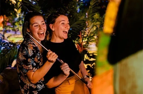 Two players laughing beside a hole, one gripping a putter, tropical planting lit warm behind them
