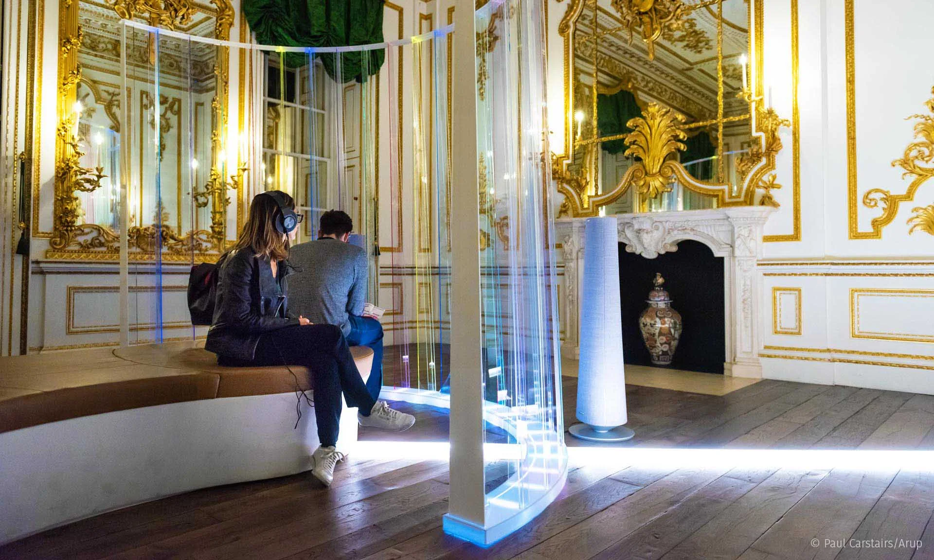 A couple sitting on the bench beside the screen, gilded chimneypiece and mirror filling the wall behind them
