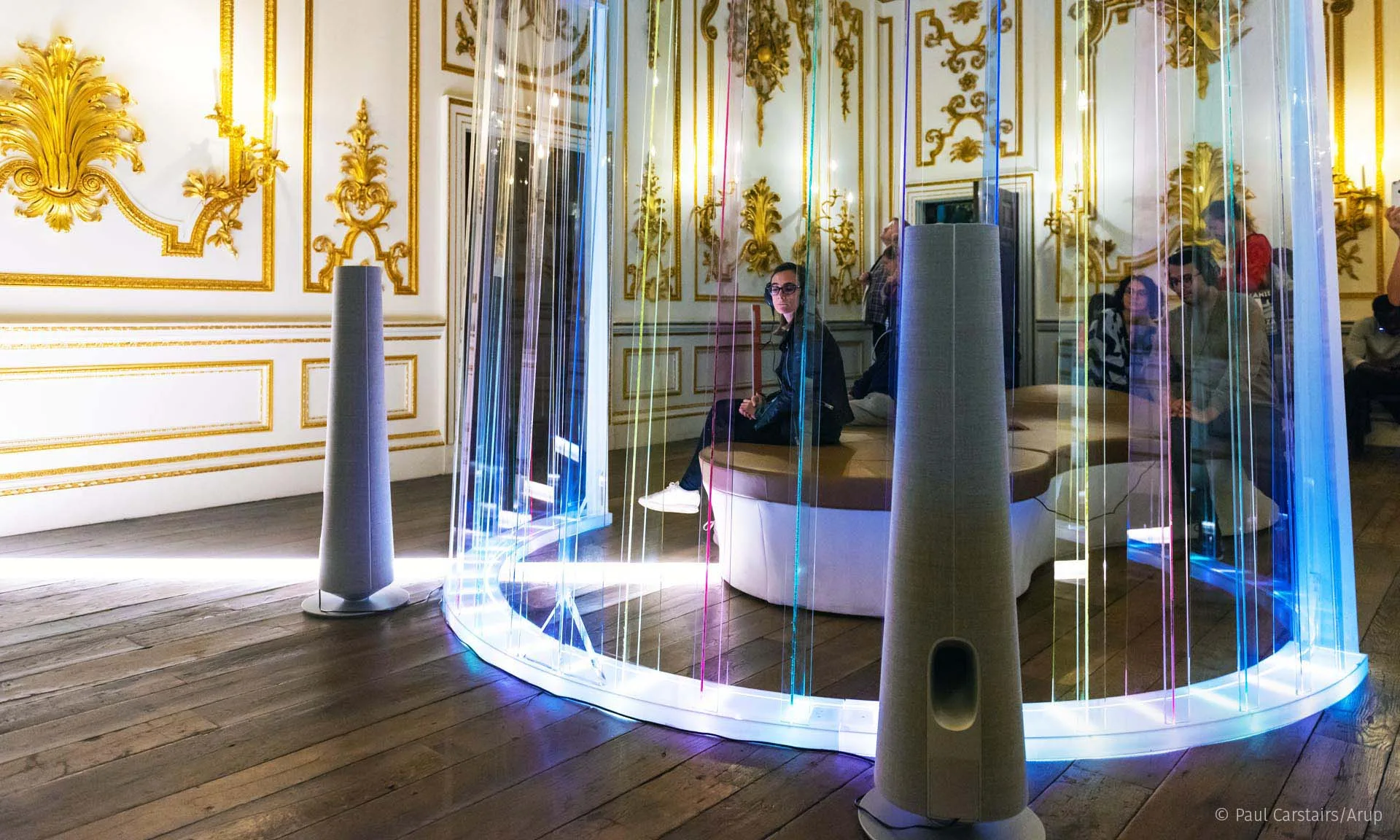 Across the room through the lit screen, a speaker tower each side and visitors seated on the bench beyond