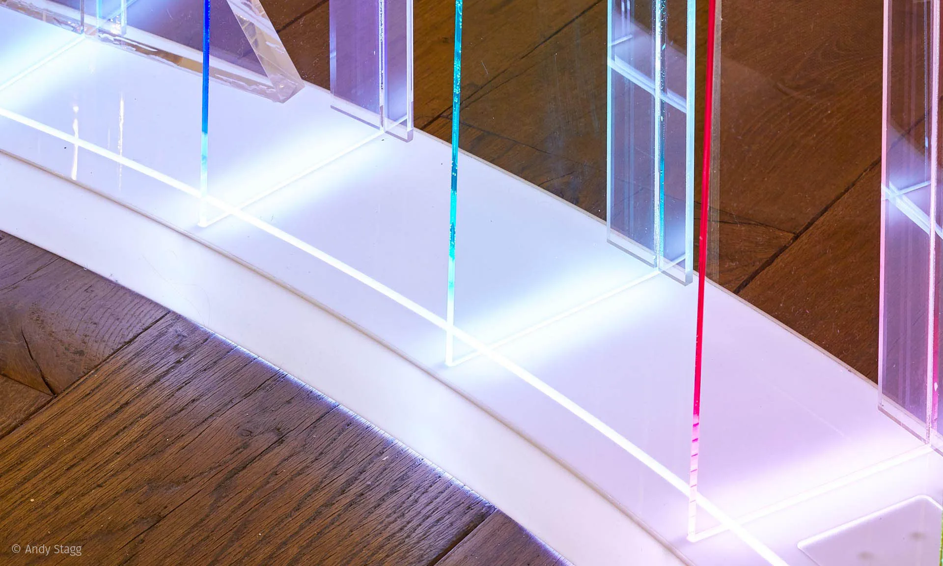 Close on the glowing base of the screen, coloured lines rising through the slat edges above the dark wooden floor
