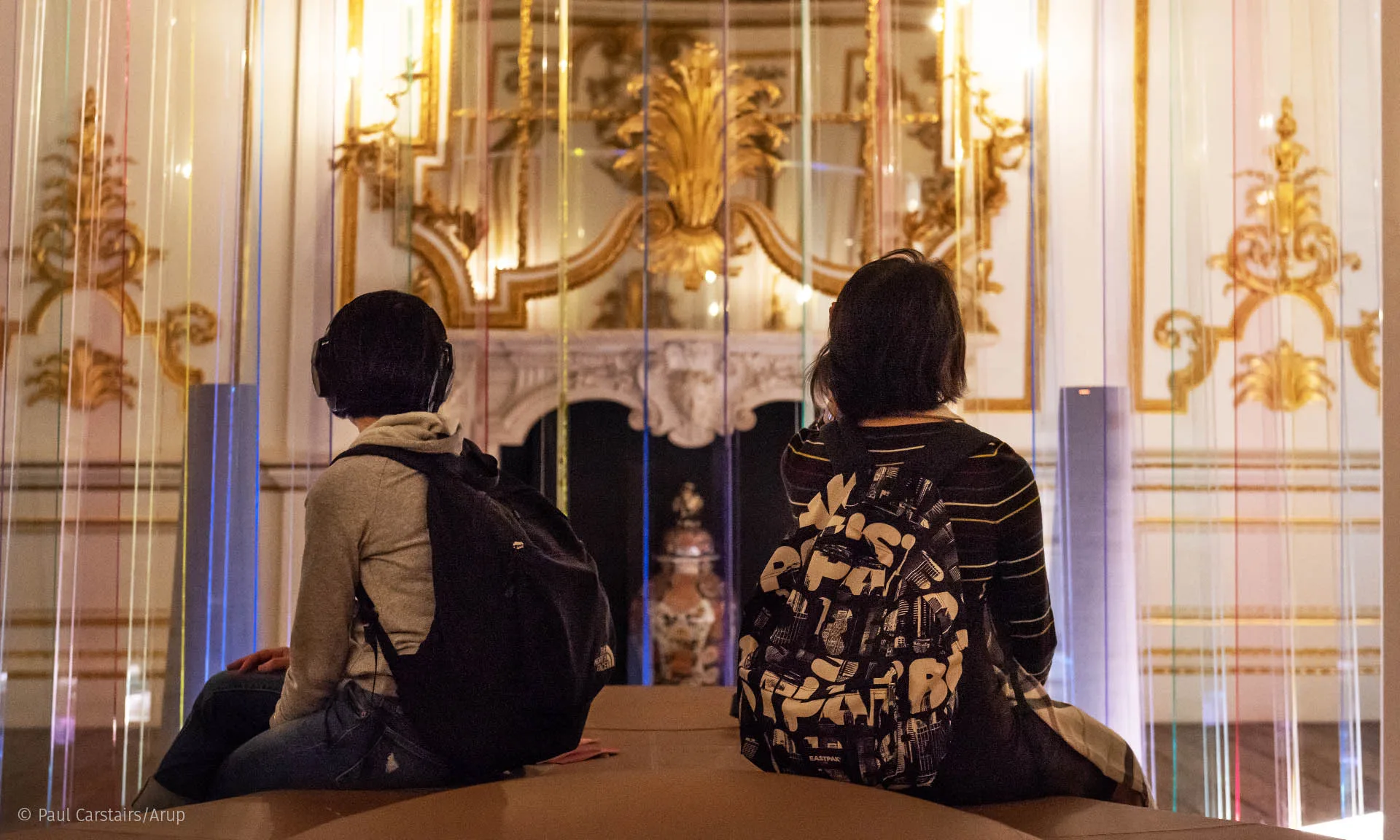 Two visitors with backpacks seated in headphones, seen through the screen looking towards the gilded fireplace