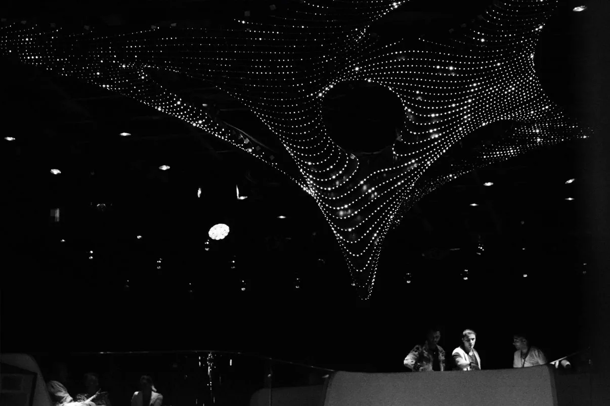 Black and white shot of the ceiling, the sculpture reading as fine arcs of white dots, staff standing at the DJ booth on the right