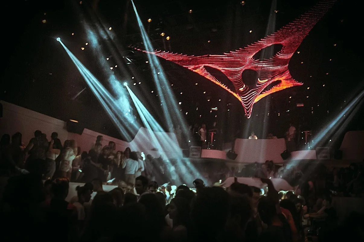 The sculpture burning orange and red while teal beams cut across the crowd below