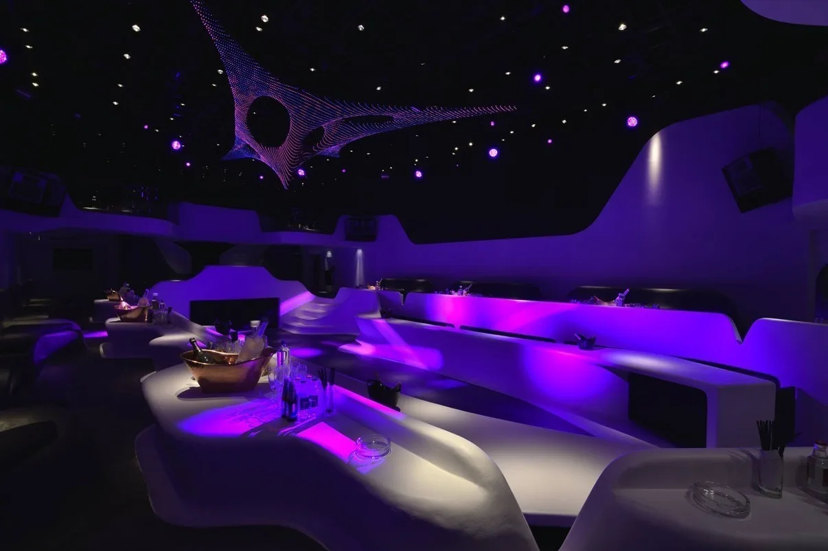 Purple and pink wash across the seating terraces, the sculpture small in the upper left and tables laid ready for service