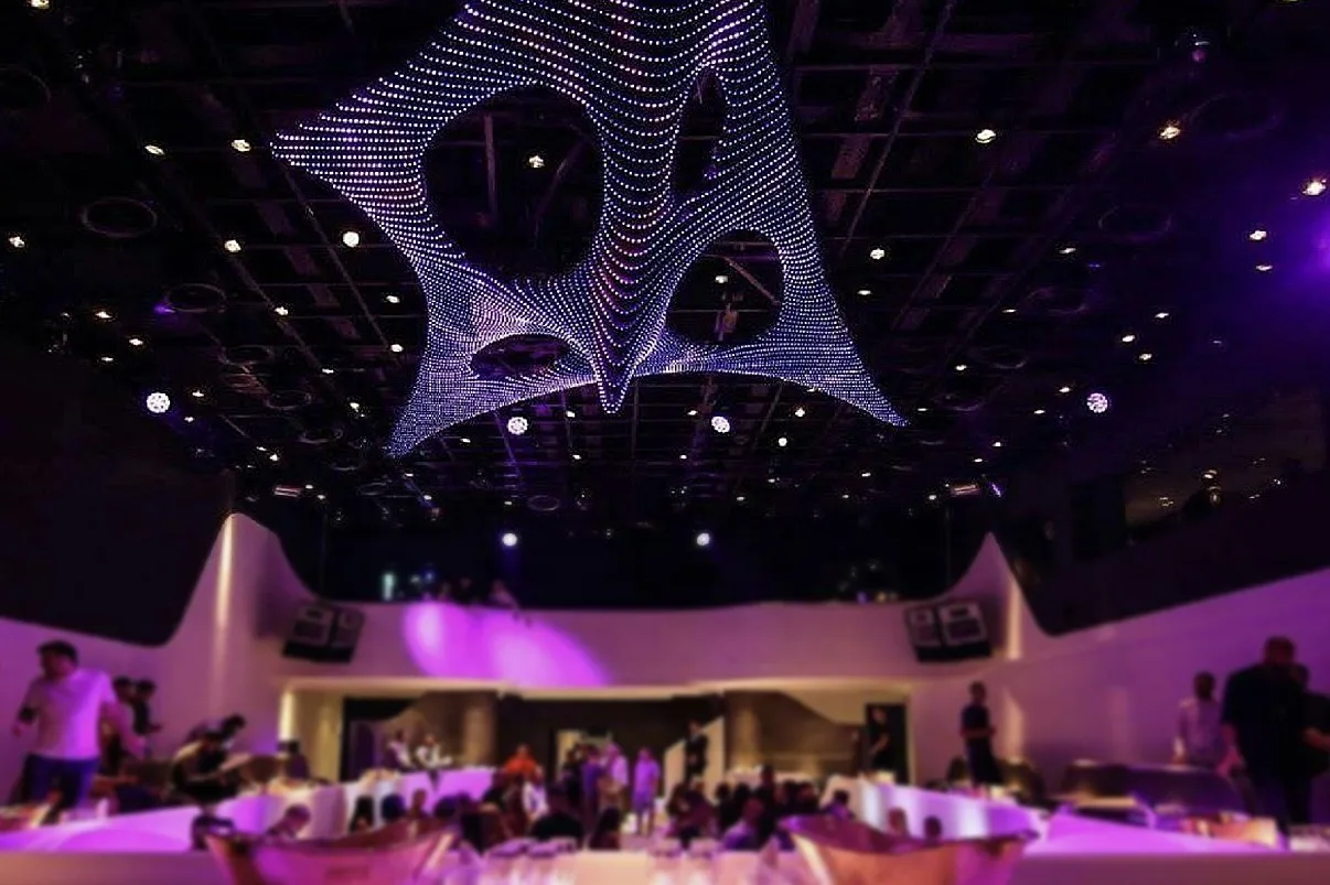 The sculpture in cool white and lilac, seen wide across a busy room with pale sculpted walls