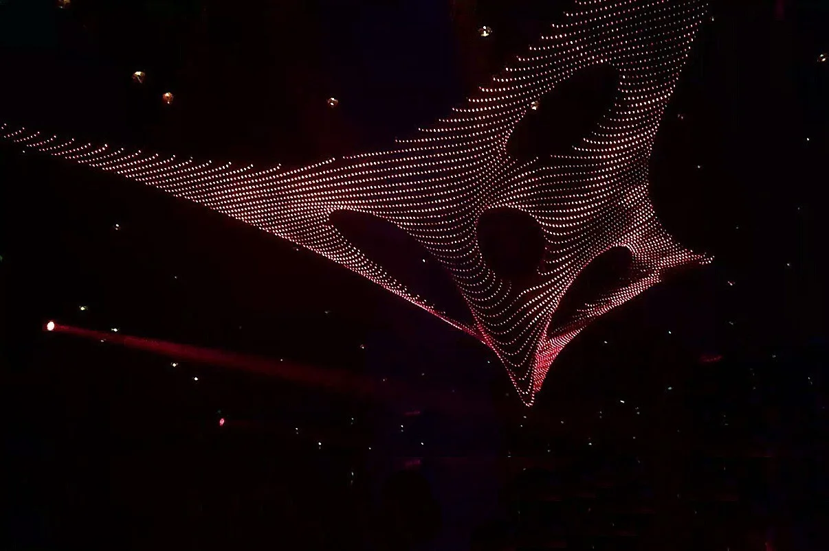 The sculpture alone against black in deep red and pink, a single beam crossing the frame from the left