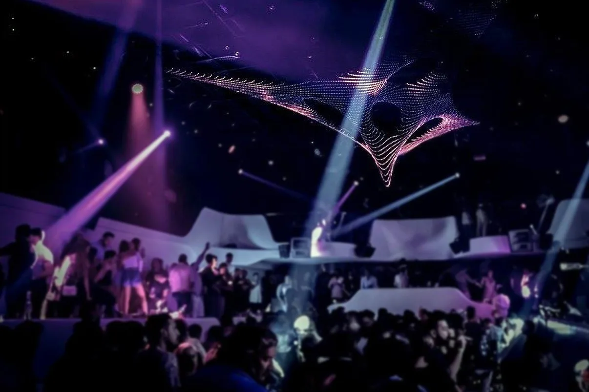White beams fanning through haze under the sculpture, arms in the air across a full dancefloor