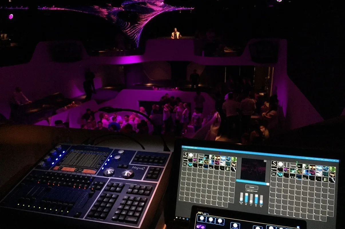 A lighting console on the balcony with the pink-lit room and dancing crowd out of focus behind it