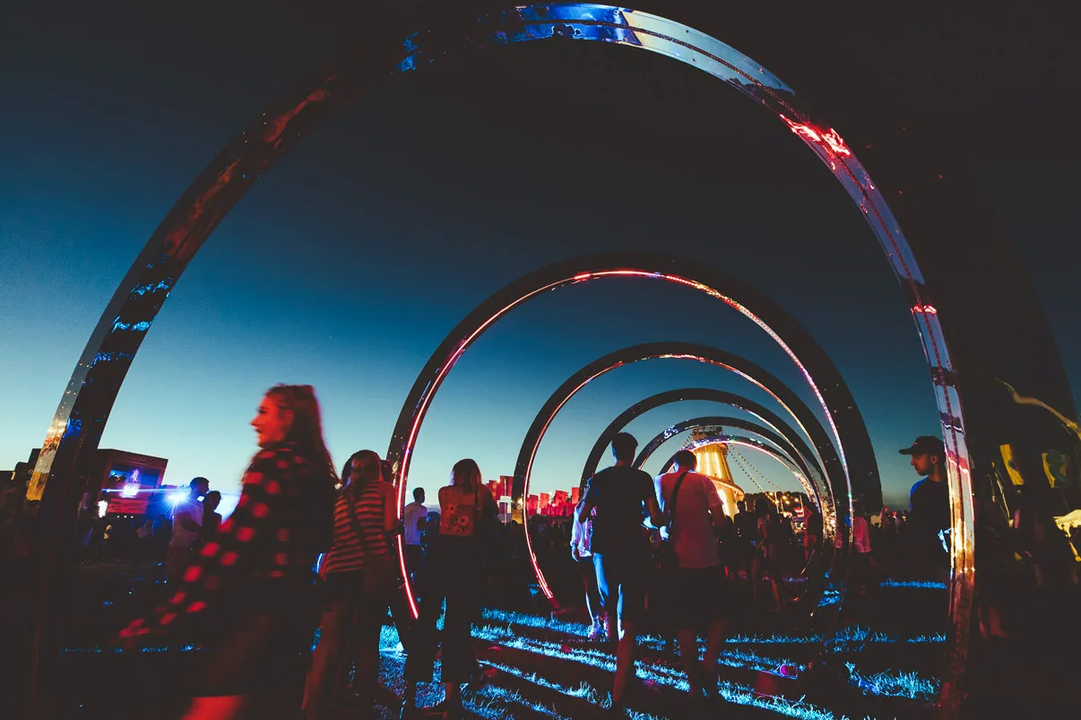 Festival goers passing through the tunnel of hoops at blue hour, red and blue light reflected in the polished steel
