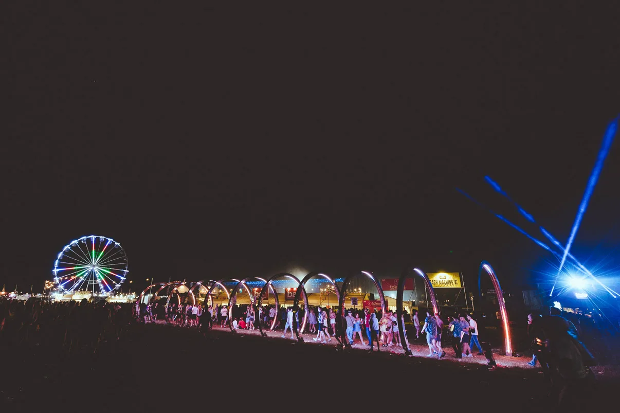 The hoop walkway seen from across the field at night, a lit ferris wheel and blue searchlight beams behind it