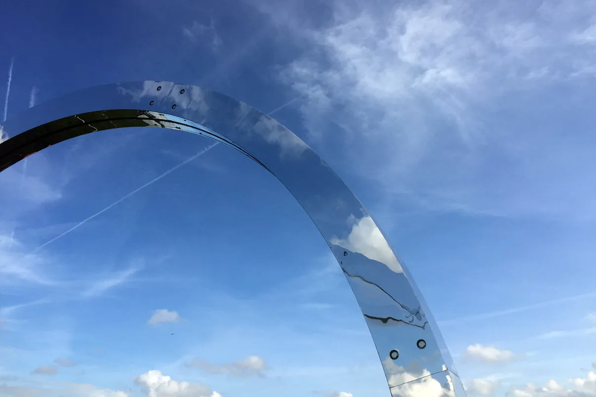 A single mirrored hoop arcing overhead, blue sky and scattered cloud reflected in the polished steel