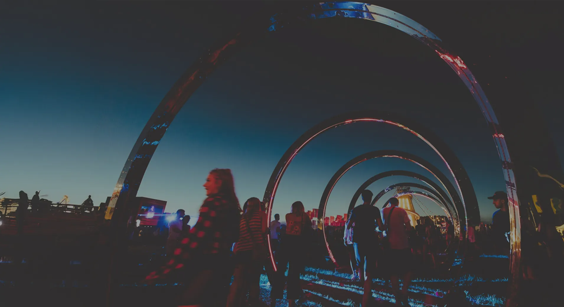 Festival-goers walking through the nested hoops of the We Are FSTVL sculpture at dusk, pixel tape lining each rim against a deep blue sky