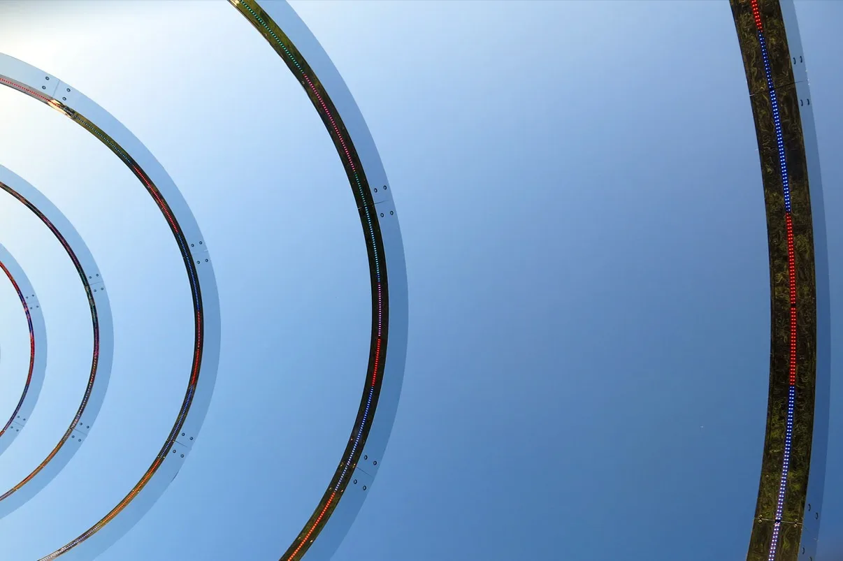 Concentric arcs of the sculpture seen from below, sunlight flaring off the polished steel against a bright sky