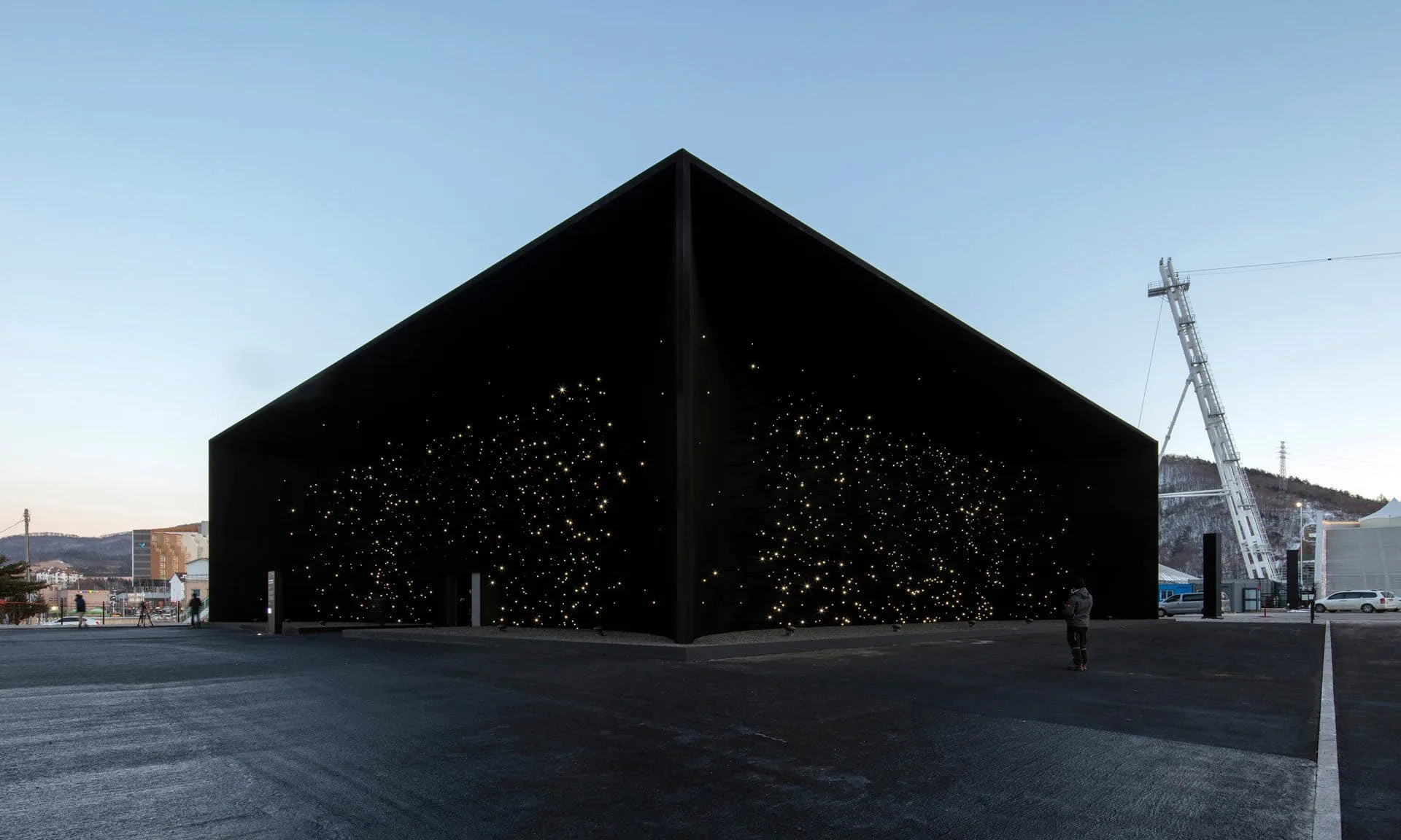 The sharp black corner of the pavilion against a pale winter sky, the plaza empty and a crane standing at the far right